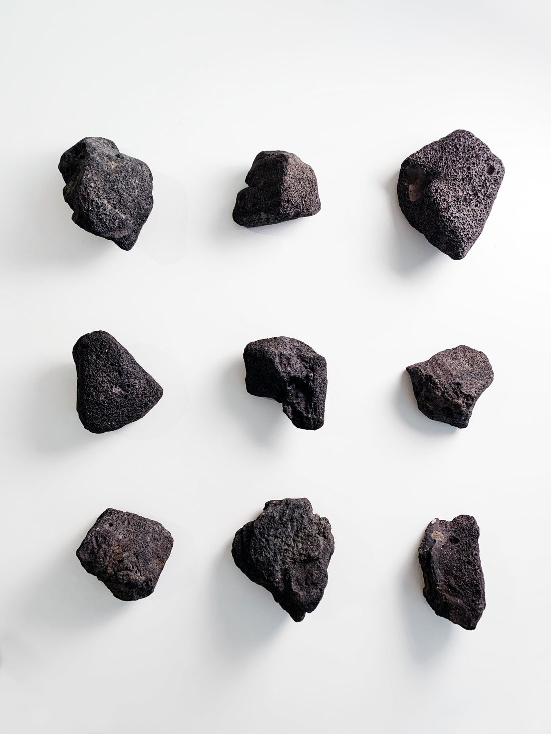 A top-down grid arrangement of nine unique, dark gray lava stones with highly textured, porous surfaces, isolated against a stark white background.