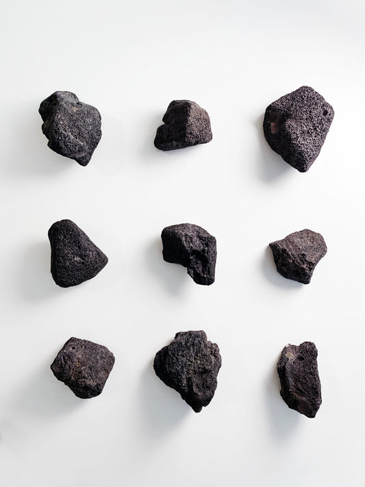 A top-down grid arrangement of nine unique, dark gray lava stones with highly textured, porous surfaces, isolated against a stark white background.