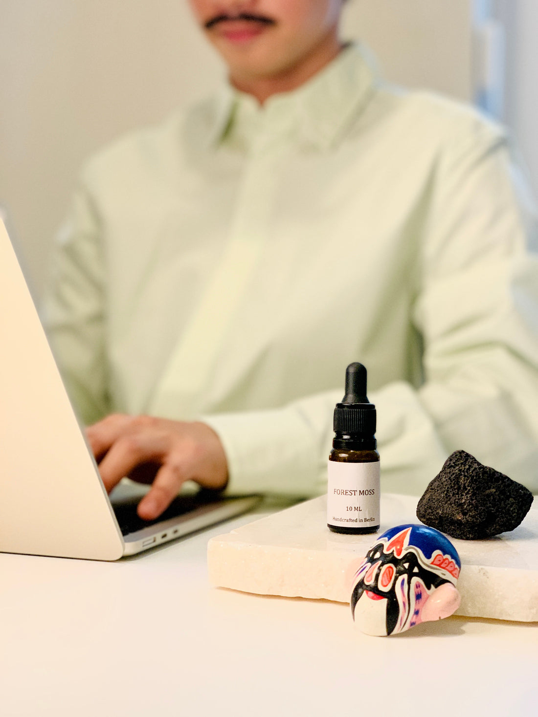 A small amber dropper bottle labeled "Forest Moss," a dark lava stone, and a small painted mask arranged on a white stone tray next to a laptop, with a blurred person typing in the background.