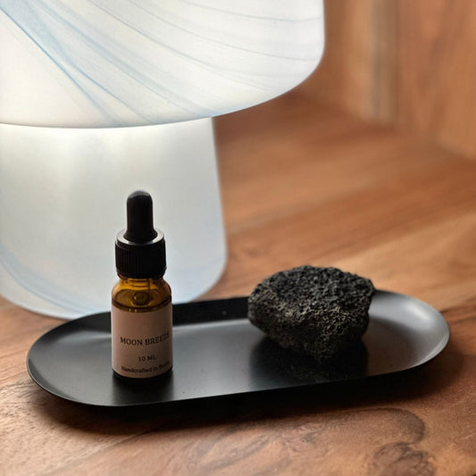 A small amber dropper bottle labeled Moon Breeze Skincare serum sits on a black tray next to a dark porous stone, with a glowing white marbled lamp and a warm wood background.